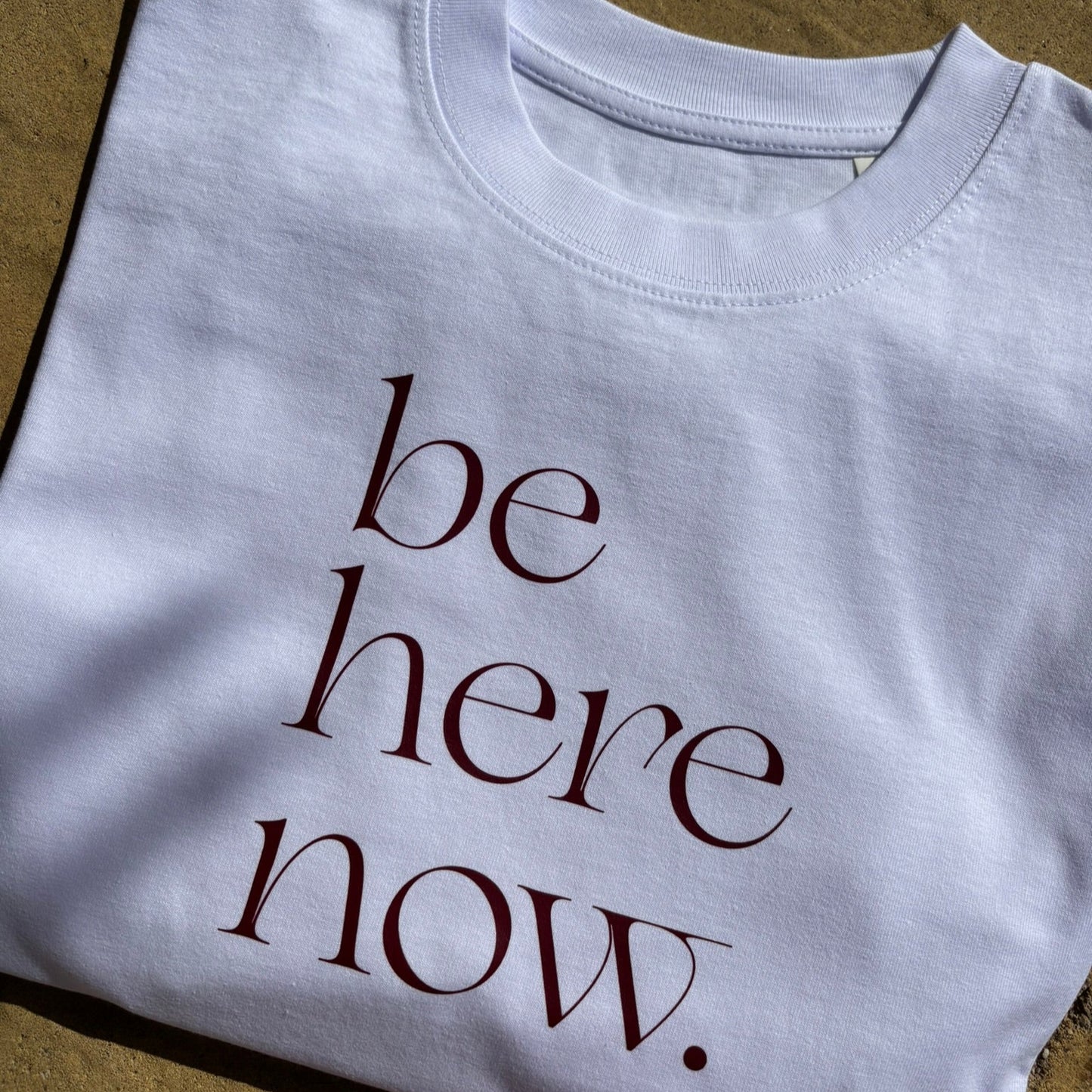 White t-shirt with 'be here now' text on a textured surface