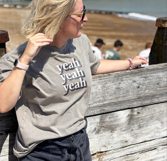 Person wearing a gray t-shirt with 'yeah yeah yeah' text, standing by a wooden railing with a beach background.