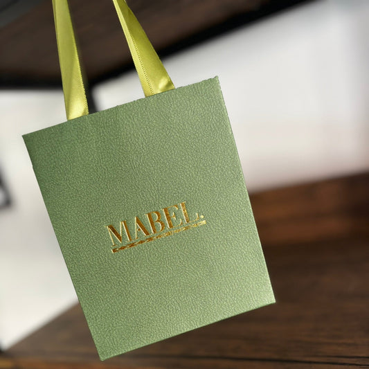 Green gift bag with 'MABEL' branding on a wooden surface