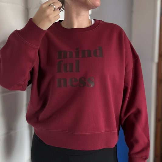 Person wearing a burgundy this is mabel boxy sweatshirt with 'mindfulness' text, standing against a white wall.