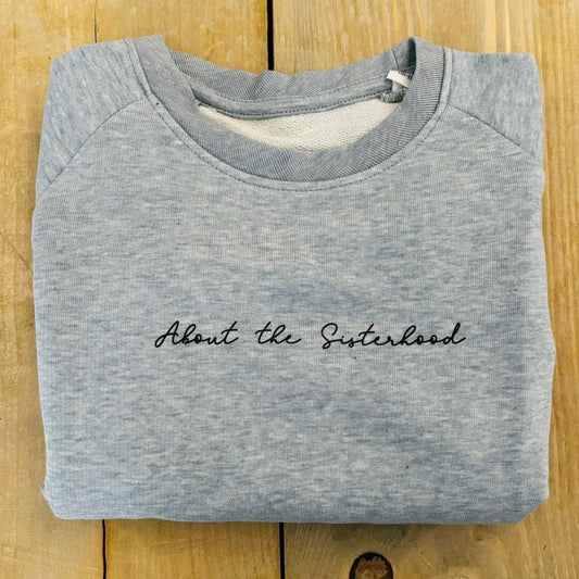 Folded heather ice blue sweatshirt with 'About the Sisterhood' text on a wooden surface