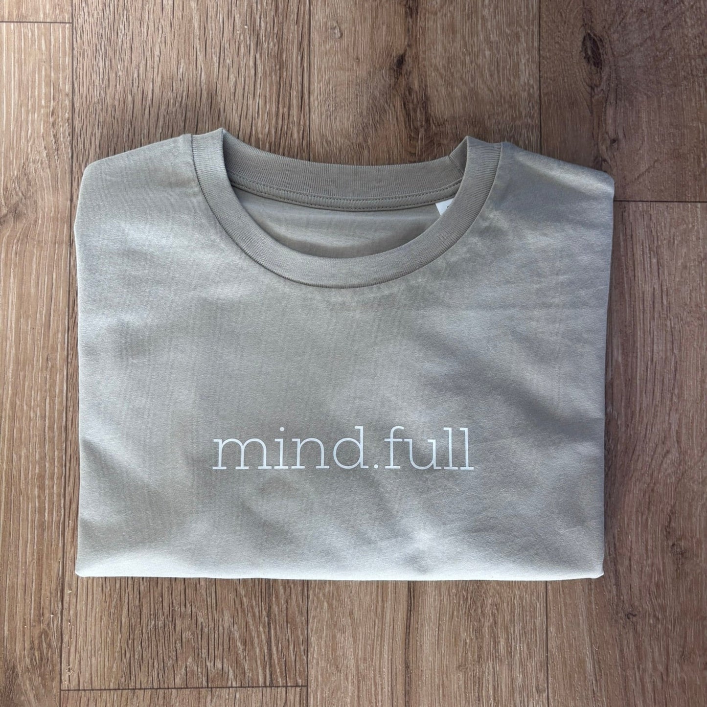 A folded stone crewneck t-shirt with 'mind.full' printed in white lowercase letters across the chest, laid flat on a brown wood plank floor.