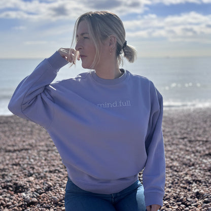 Person wearing a light purple sweatshirt with 'mindful' text on a pebbly beach.