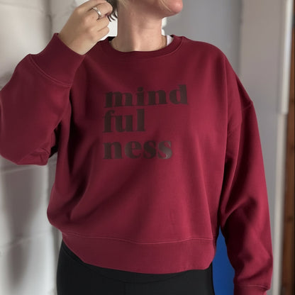 Person wearing a burgundy this is mabel boxy sweatshirt with 'mindfulness' text, standing against a white wall.