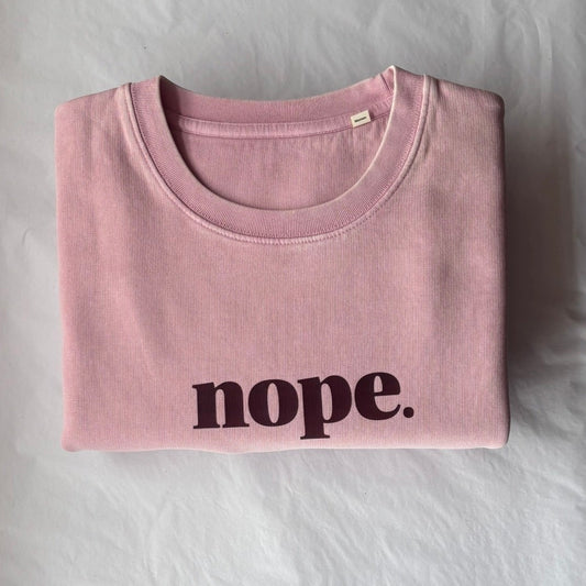 A folded light pink t-shirt with the word 'nope.' printed in black across the chest, laid flat on a white sheet of paper over a brown cardboard surface.