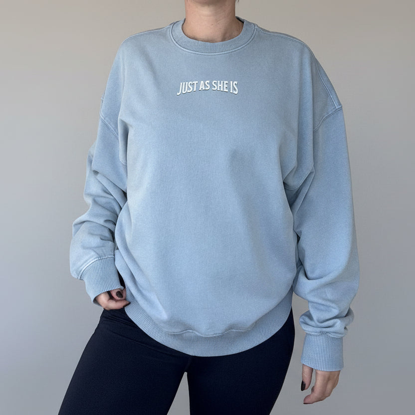Person wearing a light blue sweatshirt with 'JUST AS SHE IS' text in a kitchen setting