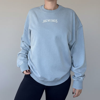 Person wearing a light blue sweatshirt with 'JUST AS SHE IS' text in a kitchen setting