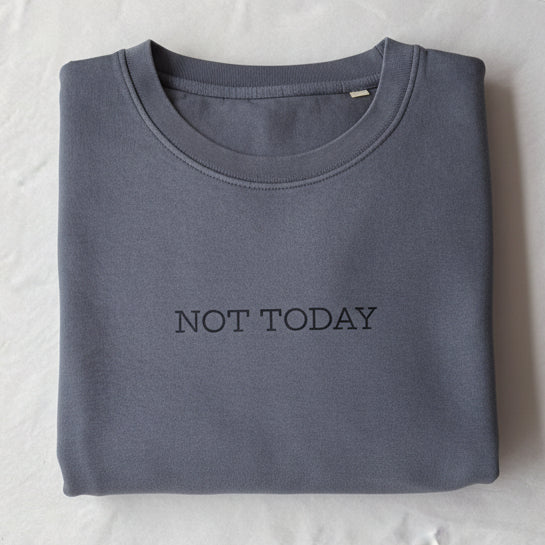 A folded slate blue crewneck sweatshirt with 'NOT TODAY' printed in black across the chest, resting on a white sheet of paper inside a brown cardboard box.