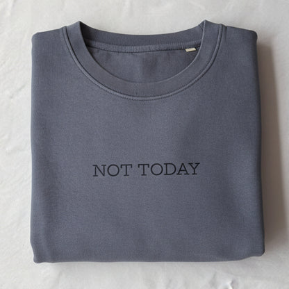 A folded slate blue crewneck sweatshirt with 'NOT TODAY' printed in black across the chest, resting on a white sheet of paper inside a brown cardboard box.