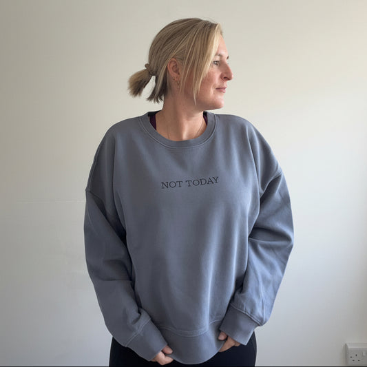 A woman with blonde hair tied back in a ponytail is wearing an relaxed fit this is mabel dusty blue-gray sweatshirt  with the text 'NOT TODAY' printed in black across the chest. 