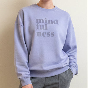 Person wearing a this is mabel oversized  light purple sweatshirt with 'mindfulness' text, sitting on a beige surface.