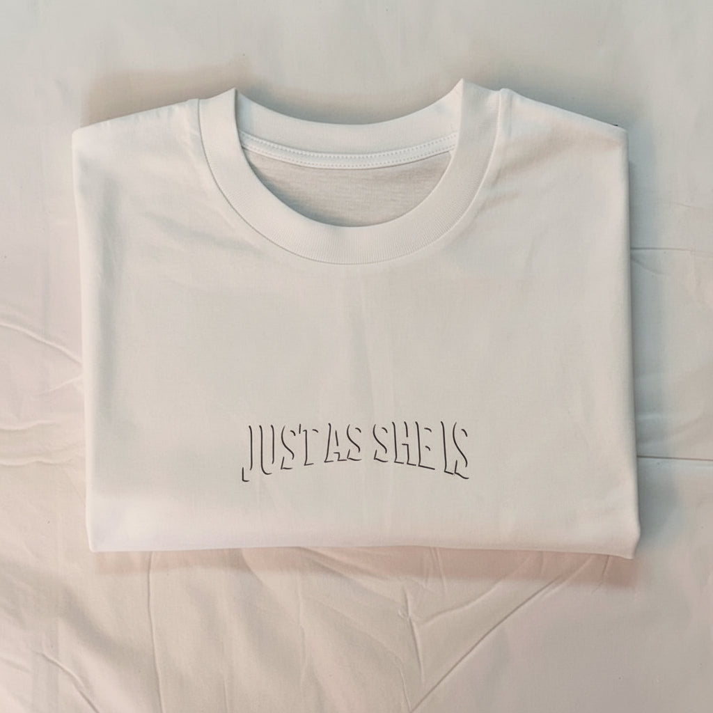 White t-shirt with 'JUST AS SHE IS' text on a light blue background