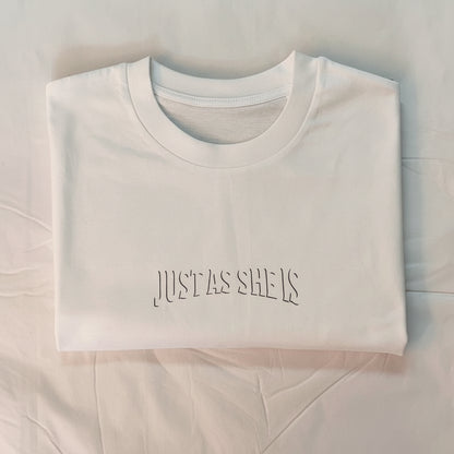 White t-shirt with 'JUST AS SHE IS' text on a light blue background