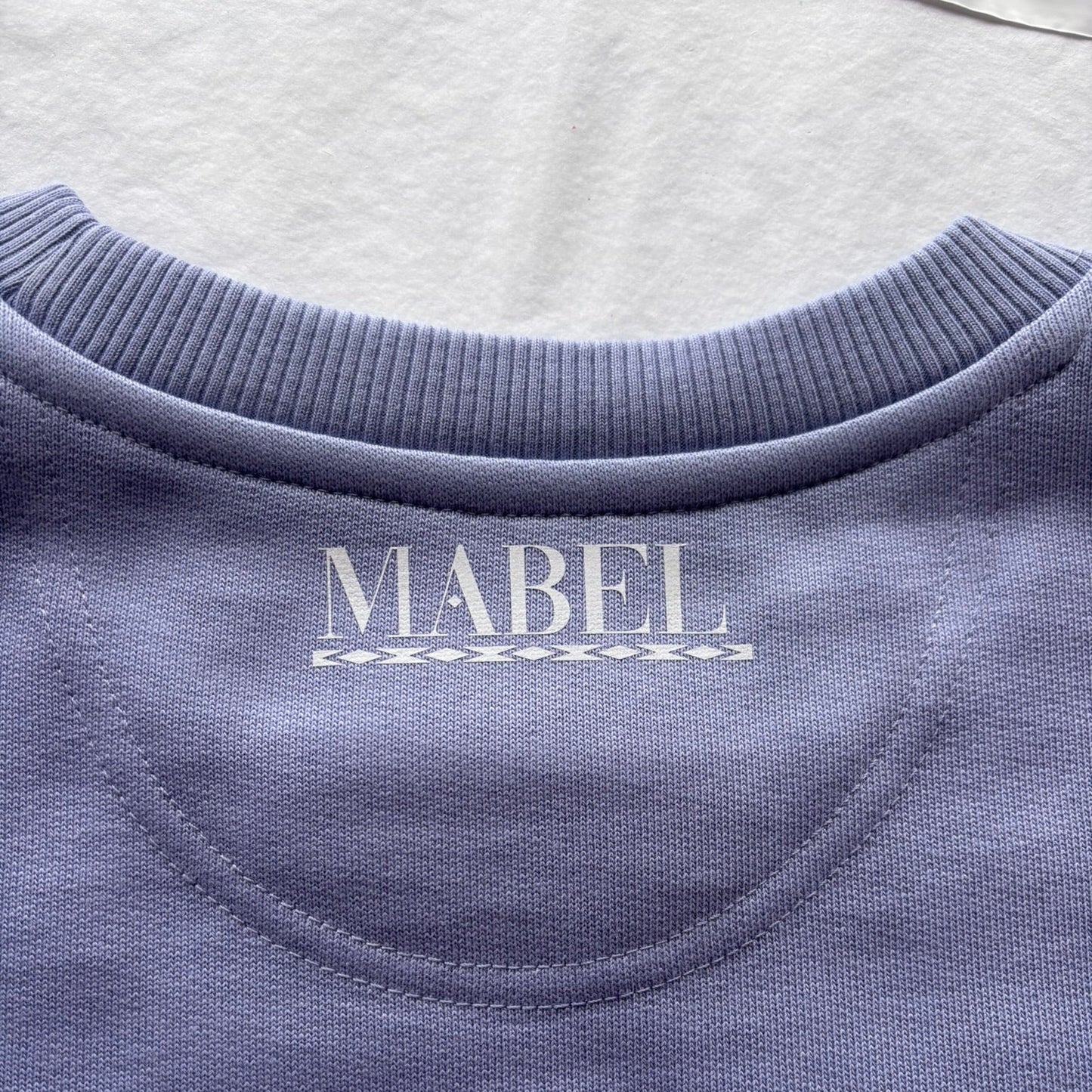 Close-up of a violet sweatshirt with 'MABEL' branding on a light grey background