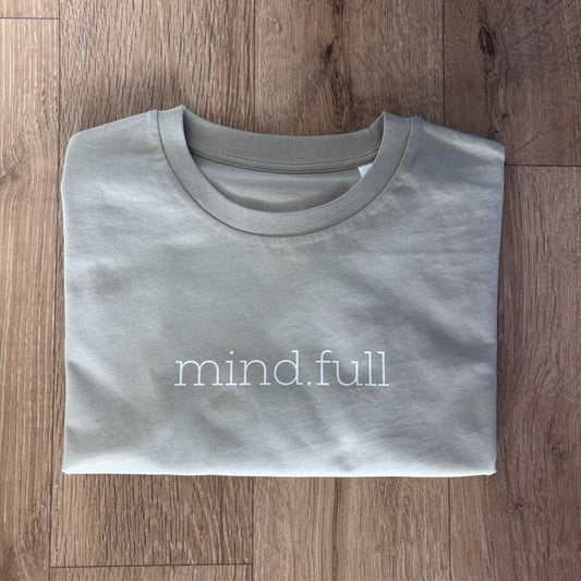 A folded stone crewneck t-shirt with 'mind.full' printed in white lowercase letters across the chest, laid flat on a brown wood plank floor.