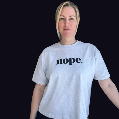 A woman with blonde hair wearing a light gray heather t-shirt with the word 'nope.' printed in navy lowercase serif letters across the chest, set against a solid black background. this is mabel 