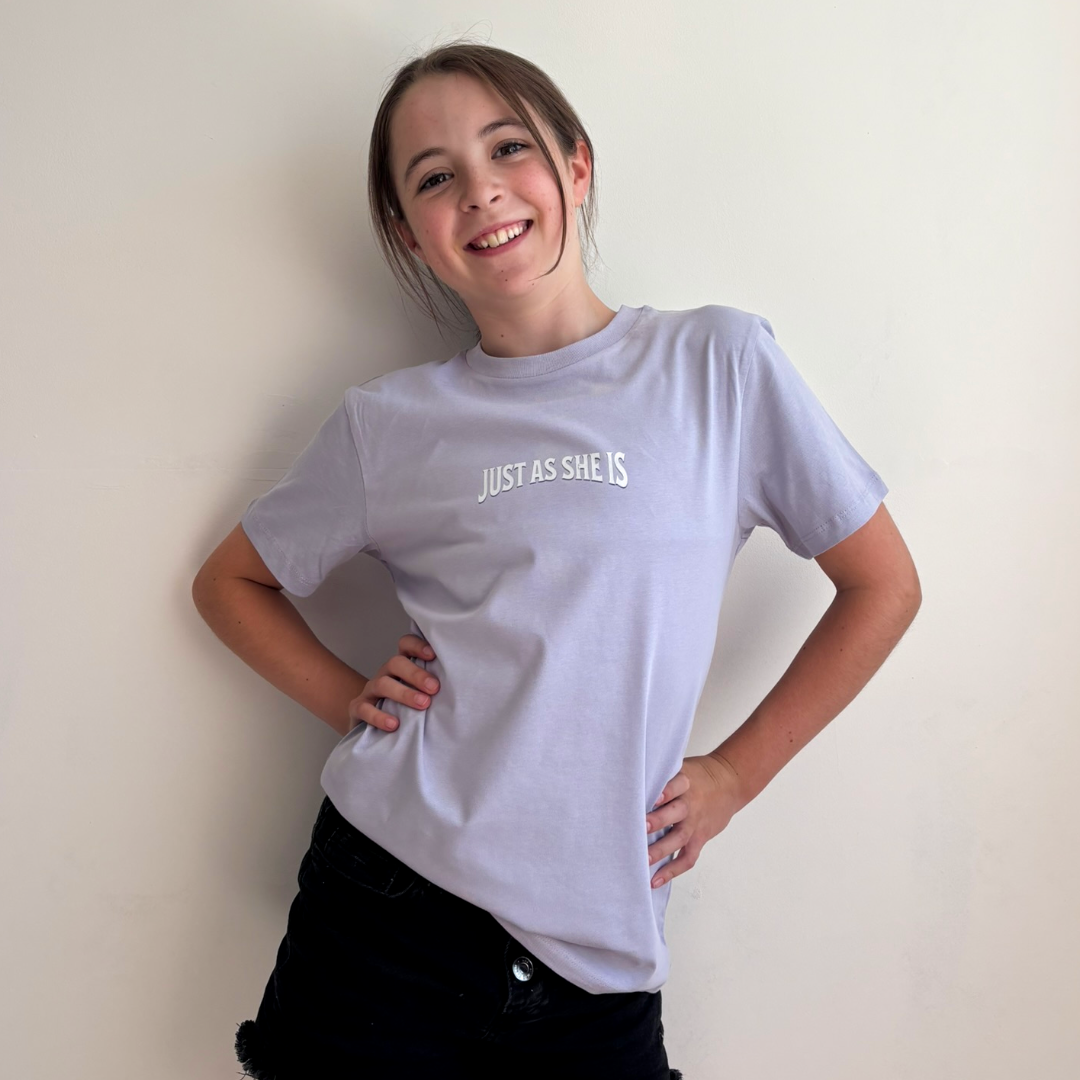 a girl smiling and posing in a light lavender t-shirt with the text 'JUST AS SHE IS' printed in white on the chest, paired with black pants, set against a plain off-white wall.