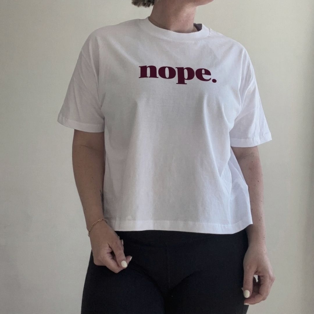 A person wearing a this is mabel this is mabel NOPE Organic Cotton Boxy T-Shirt whwhite short-sleeve cropped T-shirt with the word 'nope.' printed across the chest in dark burgundy text, paired with black pants, standing against a plain light wall.