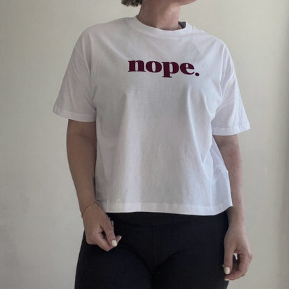 A person wearing a this is mabel this is mabel NOPE Organic Cotton Boxy T-Shirt whwhite short-sleeve cropped T-shirt with the word 'nope.' printed across the chest in dark burgundy text, paired with black pants, standing against a plain light wall.